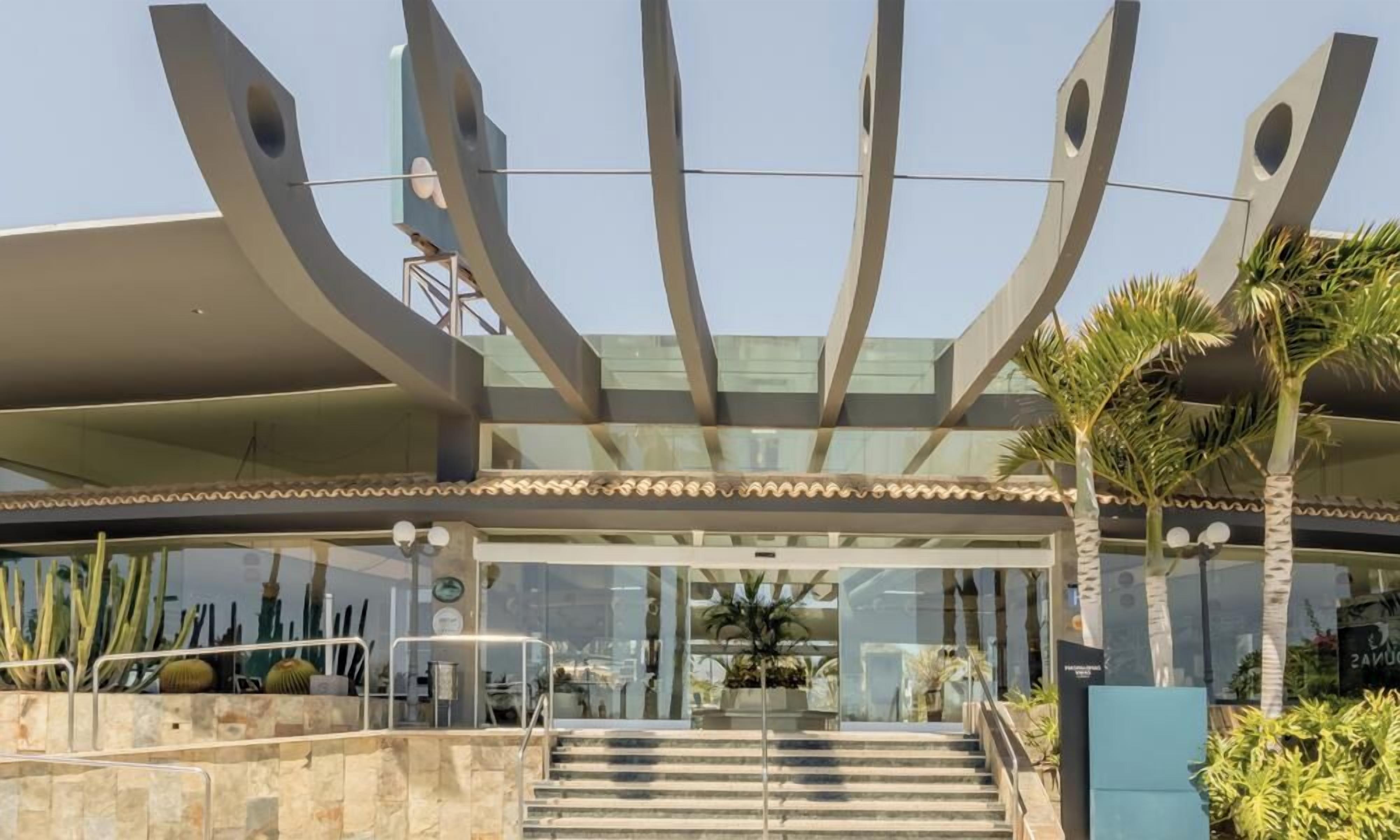 A modern building with a glass entrance, a staircase leading up to the entrance, and a palm tree in the foreground.