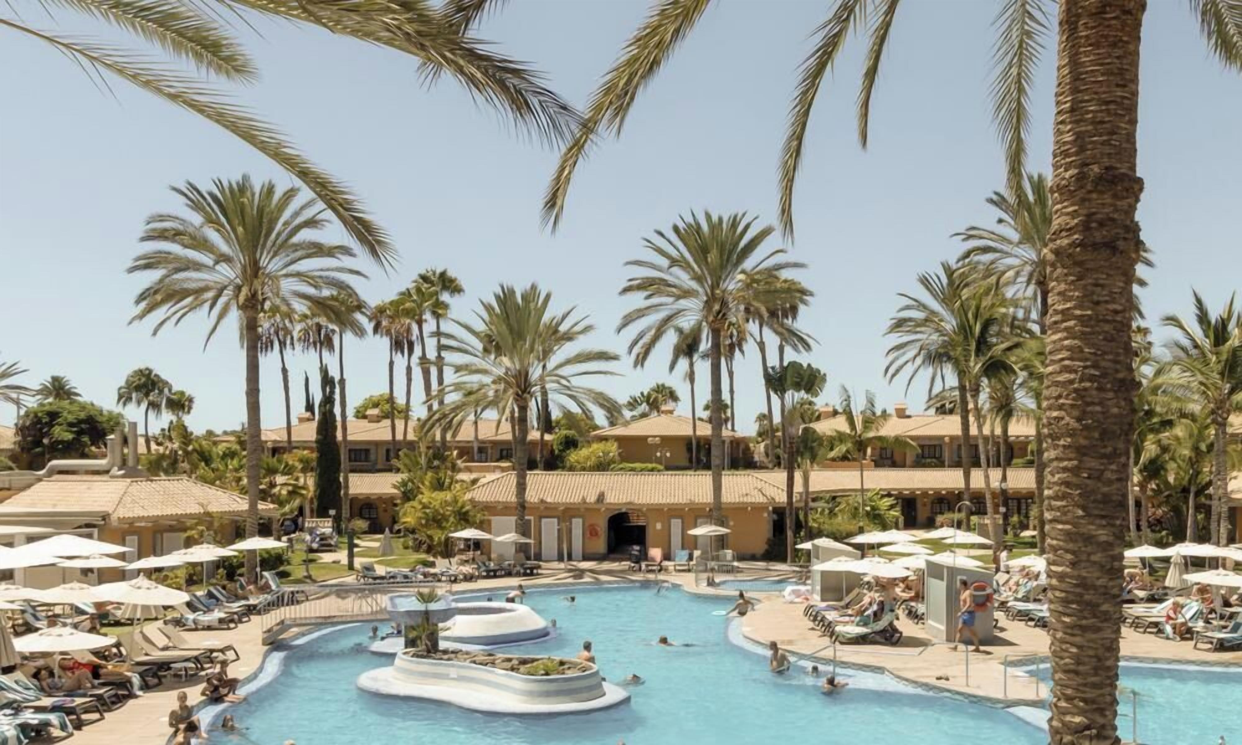 A resort with a central swimming pool surrounded by palm trees and sun loungers.