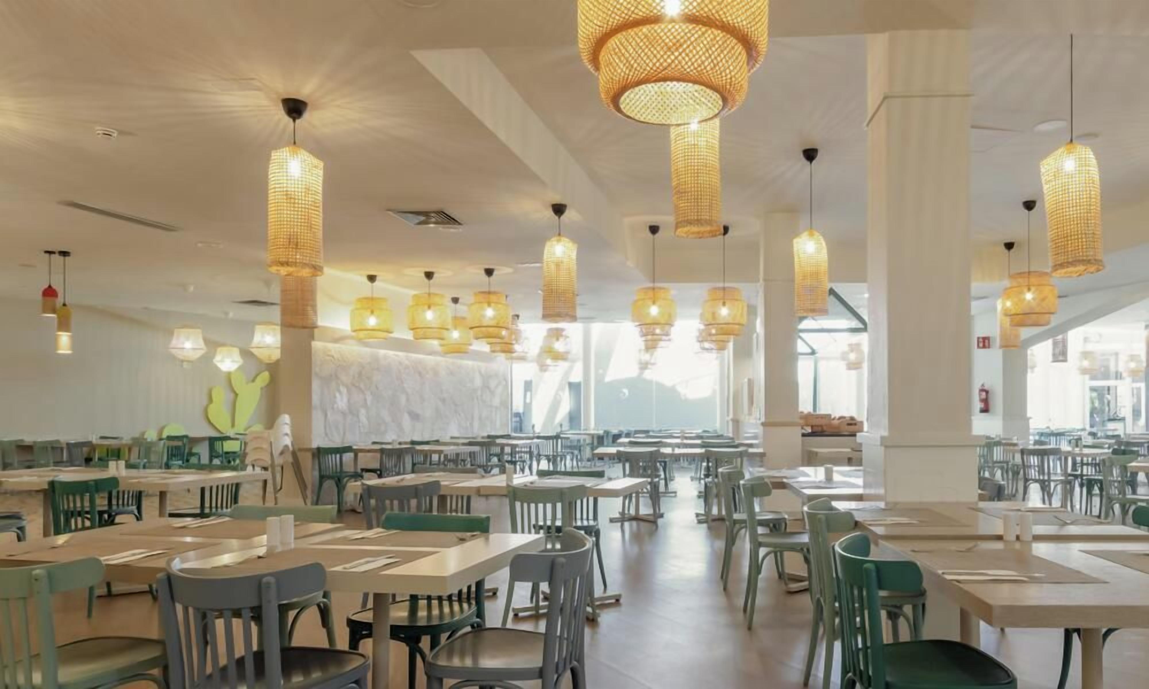 Een ruime eethoek met meerdere tafels en stoelen, uitgevoerd in een modern design met hangende rieten kroonluchters en een groot raam dat voor natuurlijk licht zorgt.