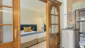 A hotel room with a wooden door, a bed with a blue and white bedspread, a nightstand, and a small desk with a trash can. - Hotiday Gran Canaria Maspalomas (San Bartolome de Tirajana)