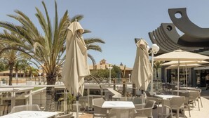 An outdoor patio area with tables and chairs, palm trees, and a modern building with a unique architectural design. - Hotiday Gran Canaria Maspalomas (San Bartolome de Tirajana)