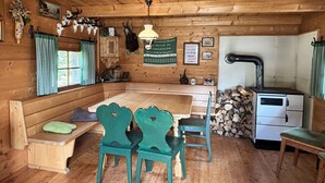 Een rustiek houten interieur met een houten bank, twee groene stoelen, een tafel, een fornuis en diverse decoraties, waaronder geweihanden en ingelijste foto's.
