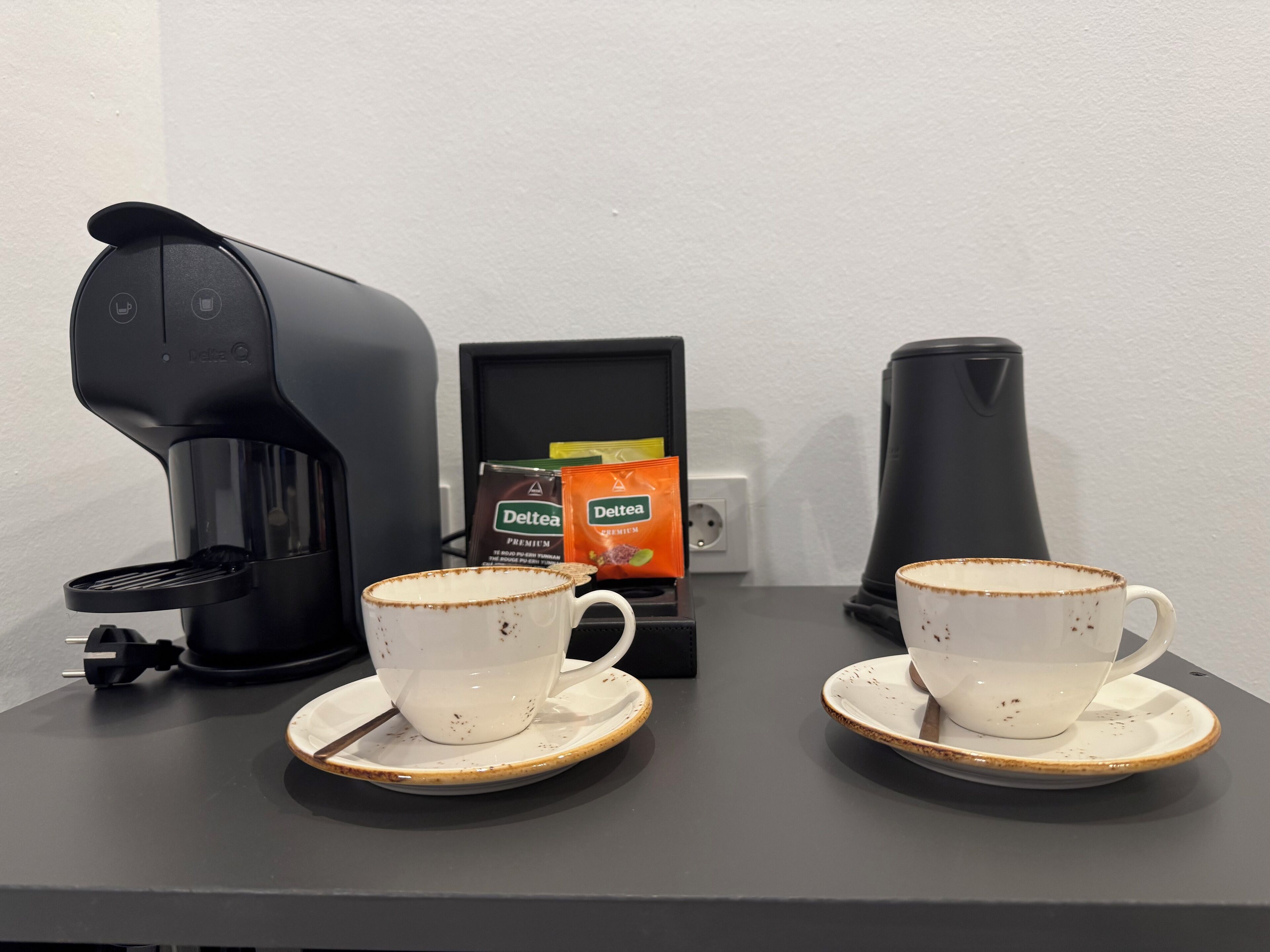 Una estació de cafè amb dues tasses blanques sobre safates, una cafetera negra i dos sobre de Deltea.