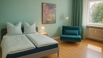 Quarto com cama, mesa de cabeceira com luminária, cadeira, janela com cortinas e uma obra de arte na parede.