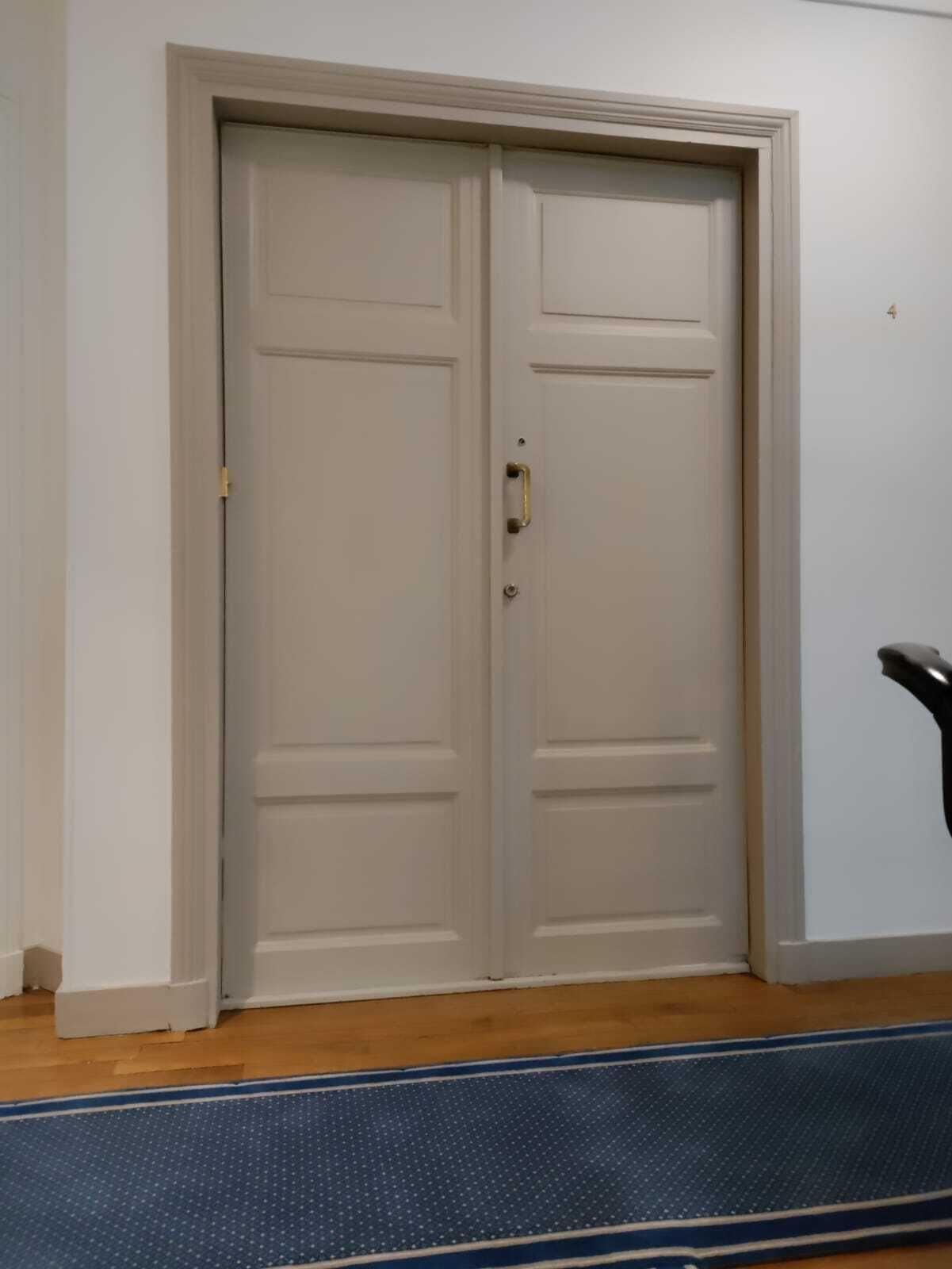 Een gesloten dubbele deur met een koperen handgreep, een houten vloer en een blauwe, geribbelde mat bij de ingang.