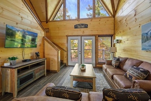 L’intérieur d’un chalet en bois, avec un téléviseur, un canapé, une table basse et un escalier menant à un balcon.