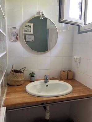 Ein Badezimmer mit rundem Spiegel, weiß gefliesten Wänden, einer Holzauskleidung, einem weißen Waschbecken, einer kleinen Topfpflanze und einem gewebten Korb.