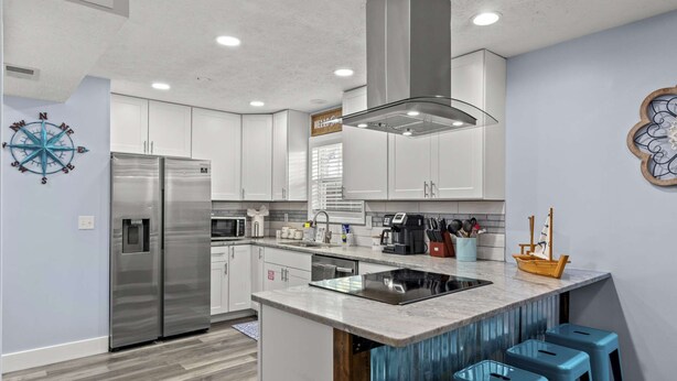 A modern kitchen with white cabinets, stainless steel appliances, a central island with stools, and decorative elements like a compass and a ship model. - Bombastic Bungalow Beach Home, just a 9 minute walk from the beach! (Panama City Beach)