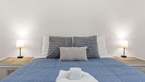A neatly made bed with a blue quilt, two gray pillows, and a white pillow. There are two bedside lamps on wooden nightstands.