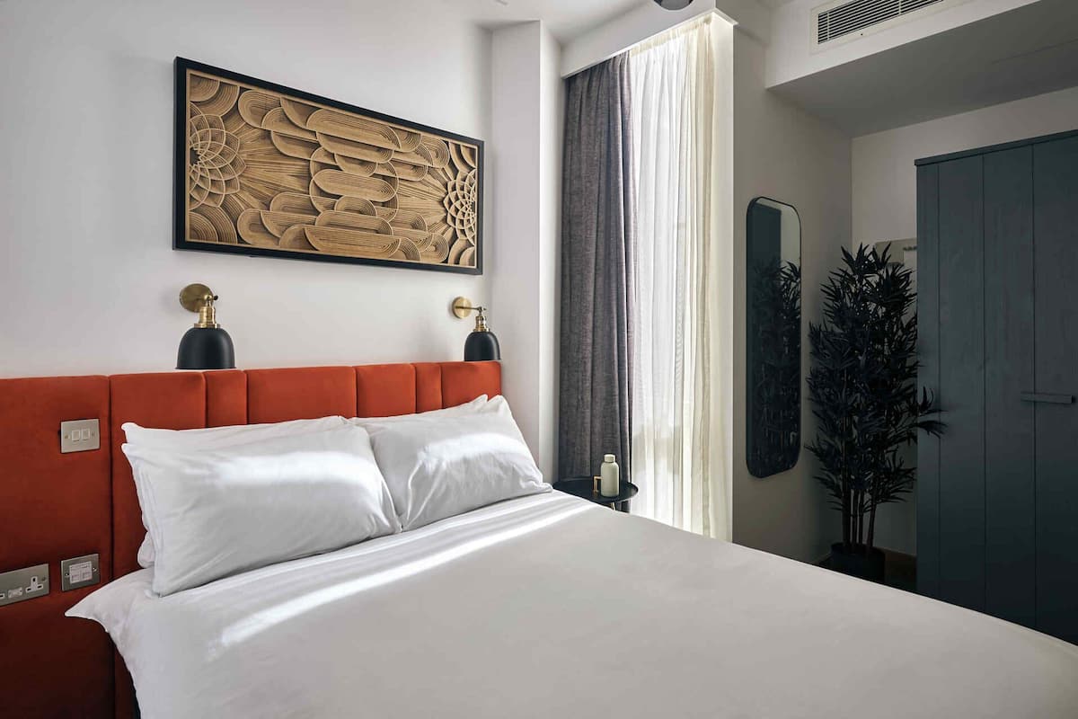 A modern hotel room with a large bed, a red upholstered headboard, a large abstract painting, a mirror, and a potted plant.
