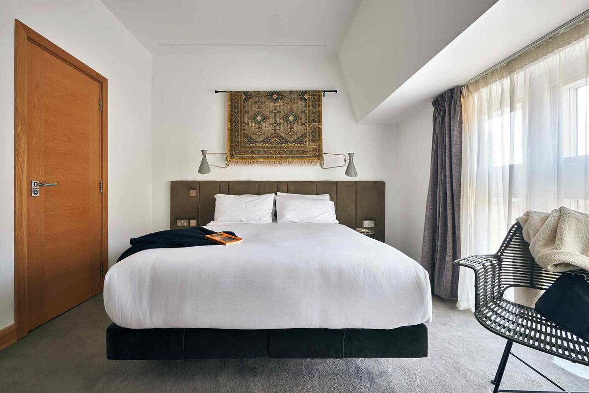 A hotel room with a large bed, a wooden door, a wall-mounted tapestry, a bedside table with a lamp, and a window with curtains.