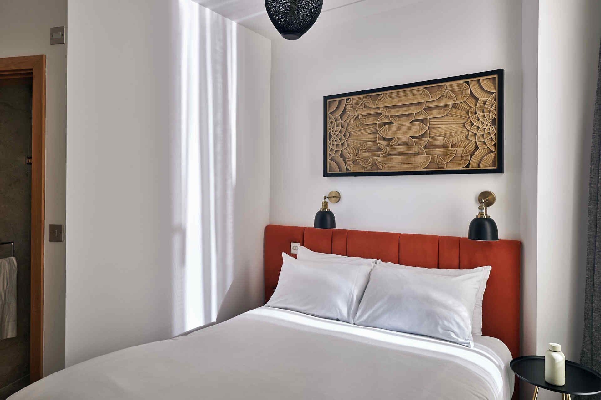 A hotel room with a large bed, a red upholstered headboard, a framed abstract artwork above the bed, and a small table with a lamp and a bottle.
