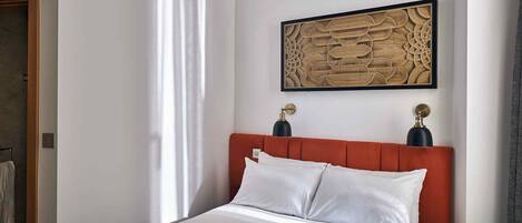 A hotel room with a large bed, a red upholstered headboard, a framed abstract artwork above the bed, and a small table with a lamp and a bottle.