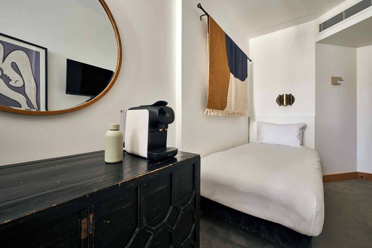 A hotel room with a bed, a bedside table with a coffee maker, a mirror, and a wall-mounted TV.
