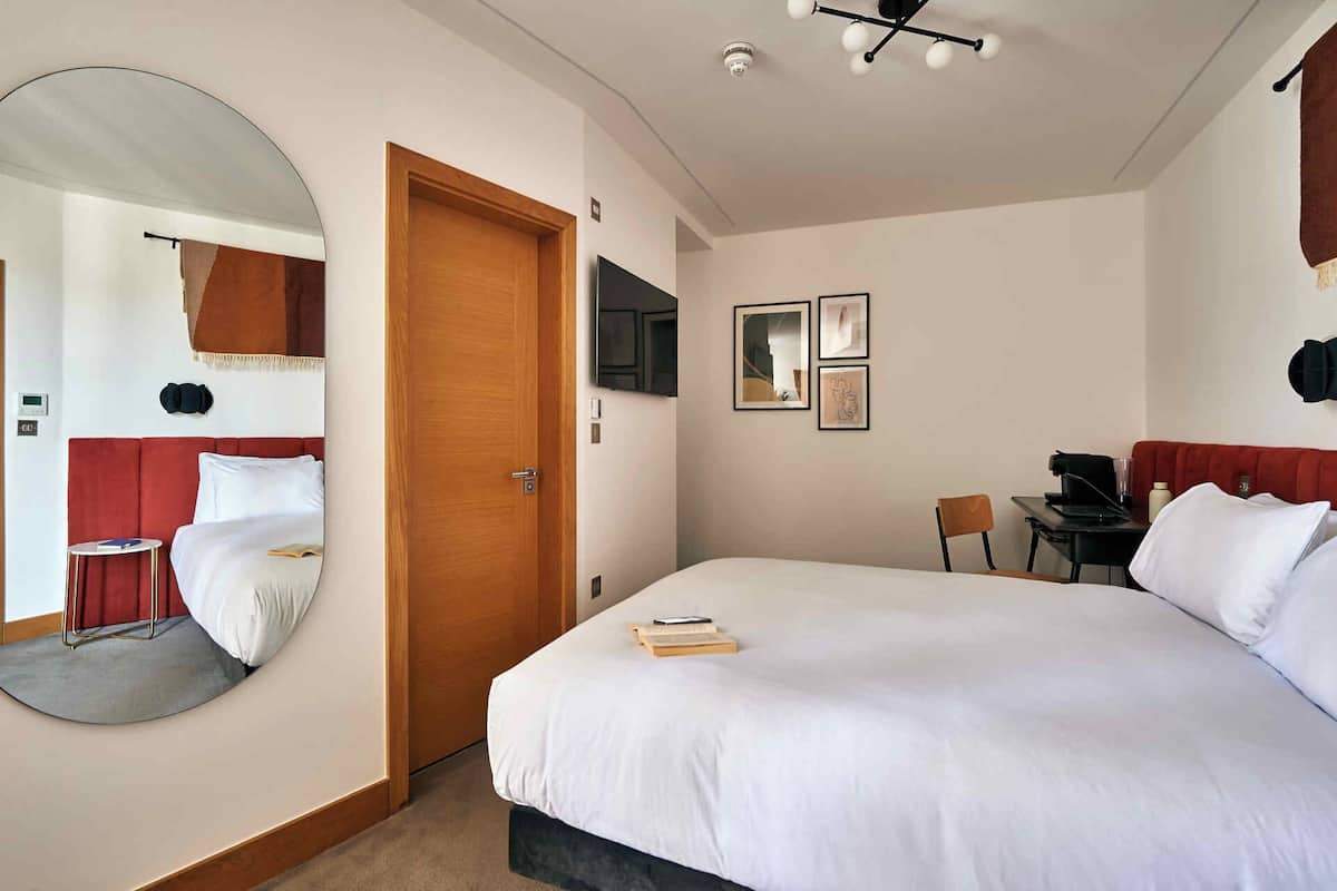 A hotel room with a large bed, a round mirror, a desk with a chair, and a small table with a lamp.