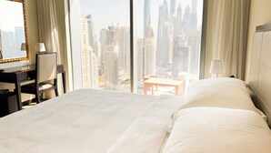 A hotel room with a large bed, a desk, and a chair. The room has a view of a city skyline through large windows.