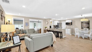 A spacious living room with a sofa, a coffee table, and a lamp. There is a kitchen area with a refrigerator, microwave, and cabinets. The room has a wooden floor and a ceiling with recessed lighting. - Tranquil Pool Home Near Siesta Key (Sarasota)