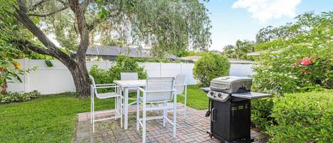 A patio with a grill, white chairs, and a tree.