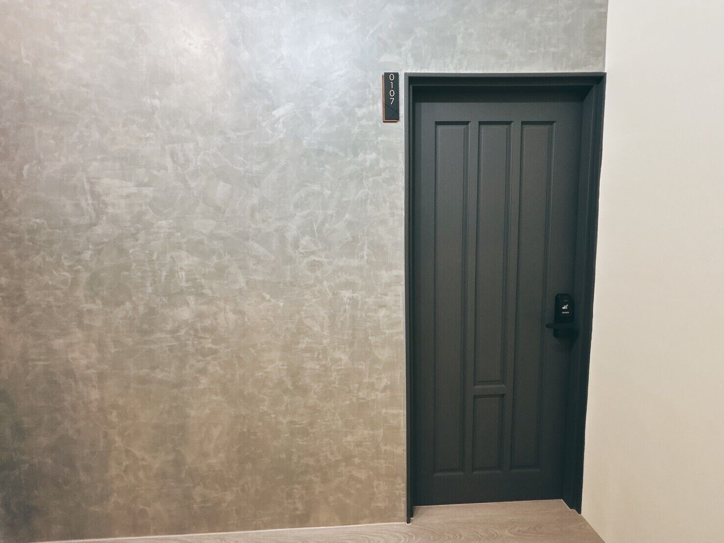 Un couloir avec un mur à texture et une porte sombre portant une plaque avec un numéro.