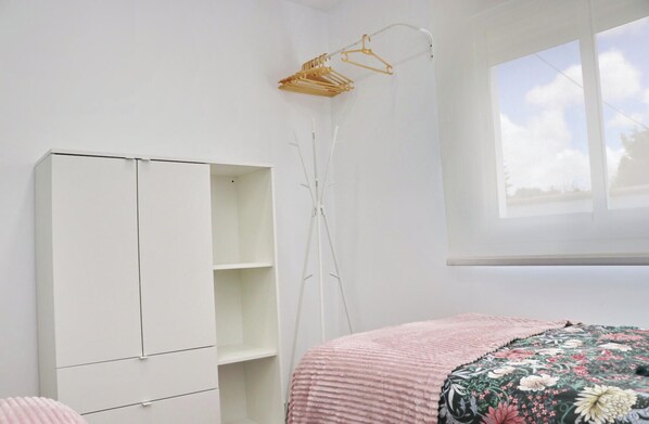A bedroom with a white wardrobe, a floor lamp, a bed with a floral bedspread, and a window with a view of the sky.
