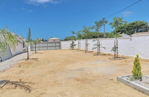 Un espace extérieur sablonneux avec un mur blanc, quelques arbres et un bâtiment en arrière-plan.