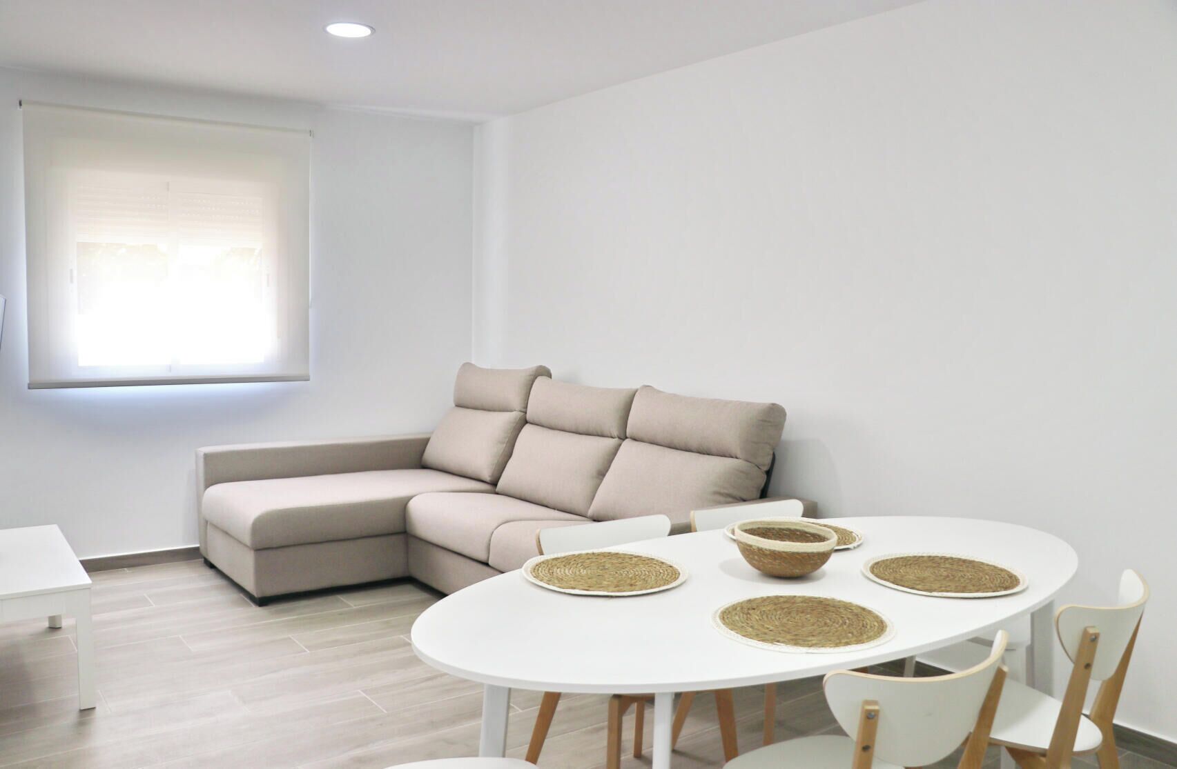 En moderne stue med en beige hjørnesofa, et rundt hvidt bord med vævede underlag og en hvid stol.