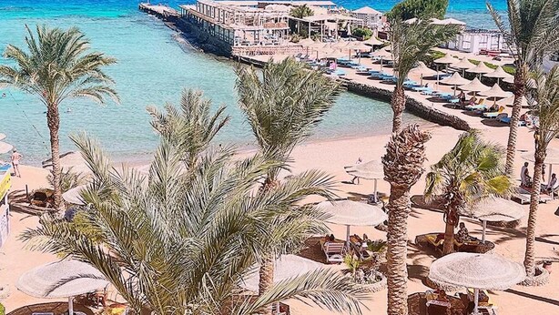 Ein Hotel am Strand mit einer Pier, die sich ins Meer erstreckt, Palmen und Liegestühlen.