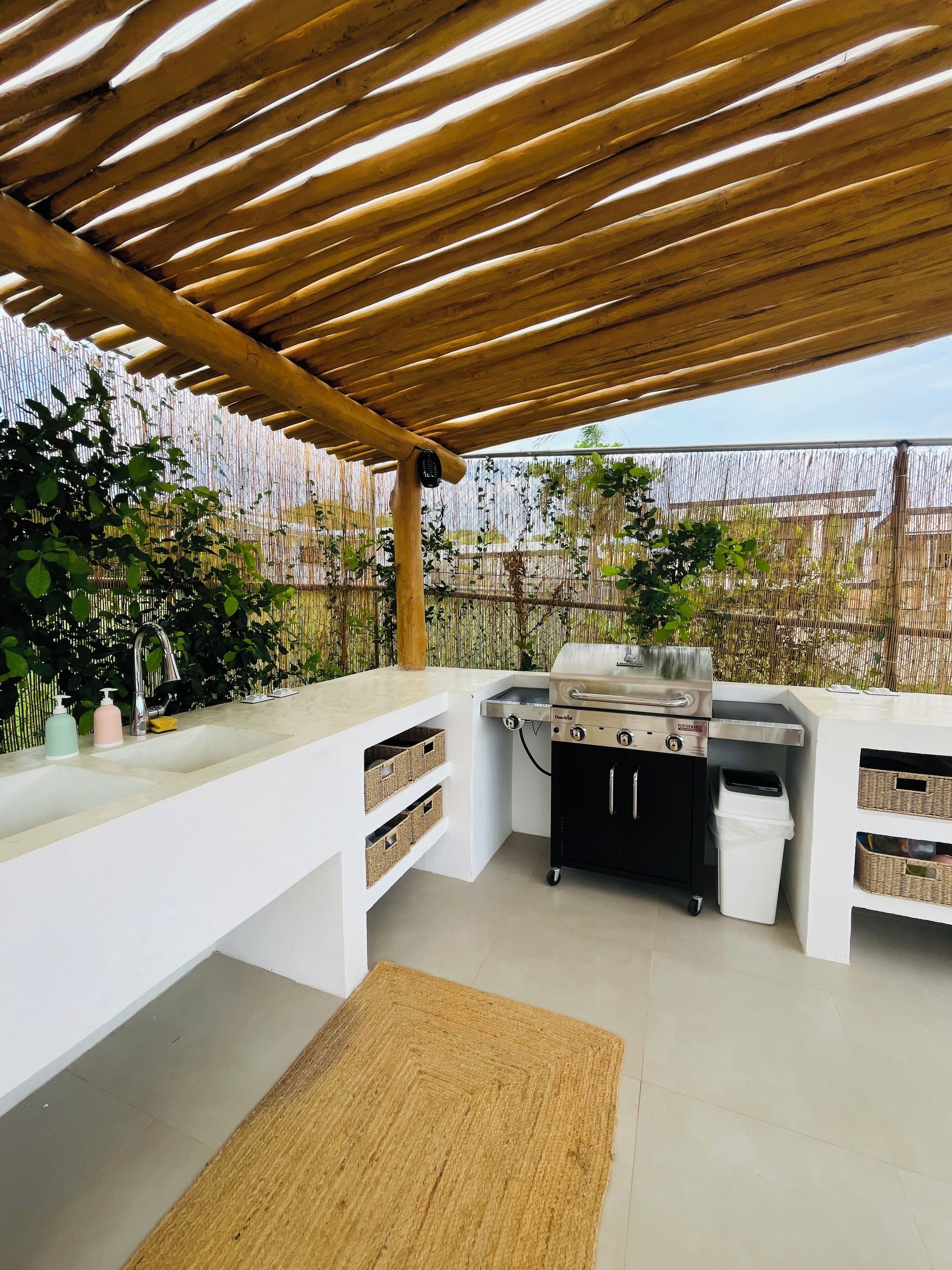 Cozinha externa com churrasqueira, armários brancos e pérgola de bambu.