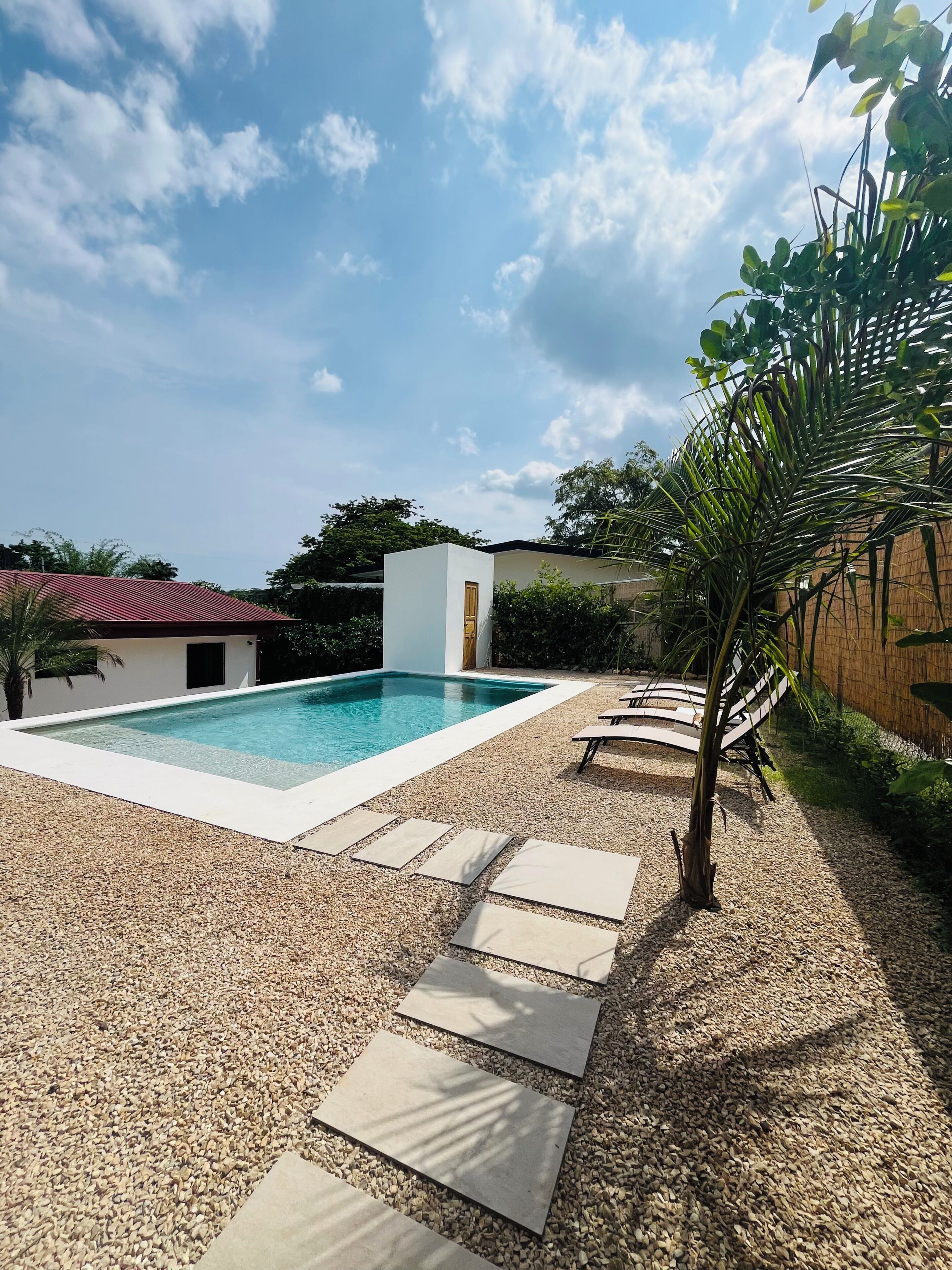A swimming pool with a white deck, surrounded by pebbles and a few lounge chairs.