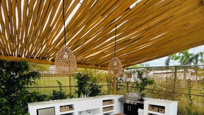 Een buitenkeuken met een houten tafel en witte stoelen, een hanglamp en een witte opbergkast met manden.