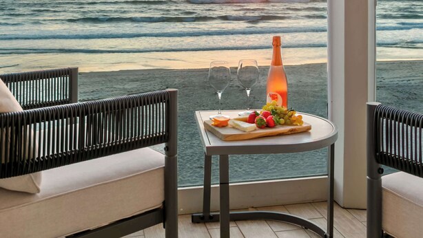 A balcony with a view of the sunset over the ocean, a table with a bottle and fruit, and two chairs.