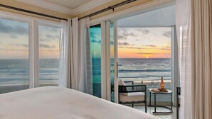 A hotel room with a large bed, a chair, a small table with a bottle and glasses, and a view of the ocean at sunset.