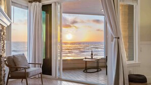 A room with a view of the ocean at sunset, a chair, a small table with a bottle and glasses, and a stone wall.