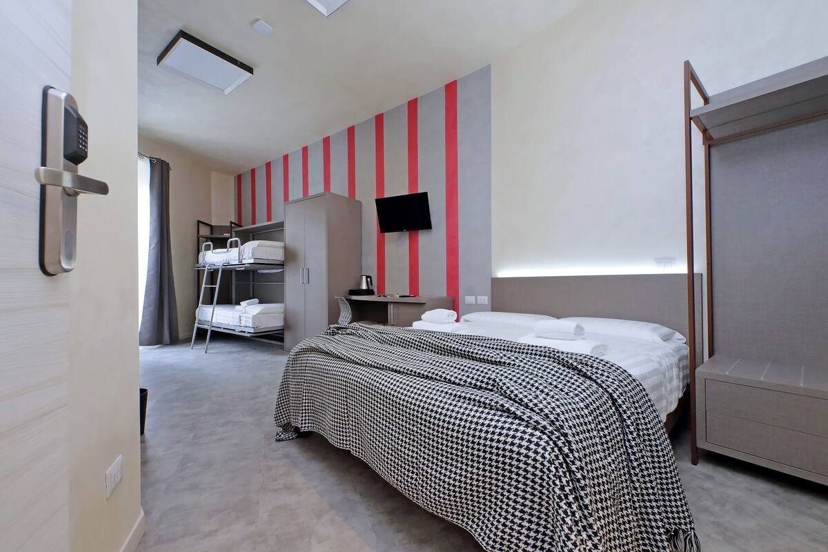 A hotel room with a bed, a TV mounted on the wall, a wardrobe, and a desk with a chair.