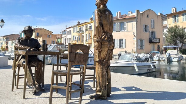 Una statua di un uomo con cappello e cappotto in piedi accanto a un tavolo e due sedie.