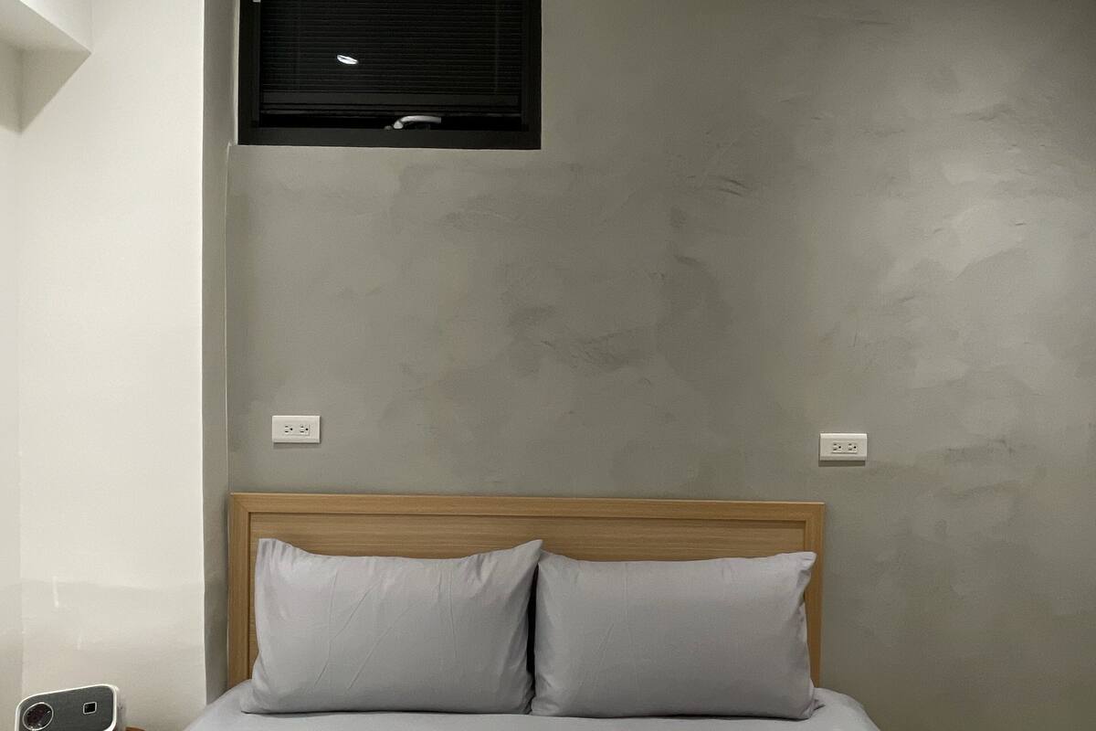 Quarto com cama, duas almofadas, cabeceira de madeira, mesa de cabeceira com abajur e ar-condicionado fixado na parede.