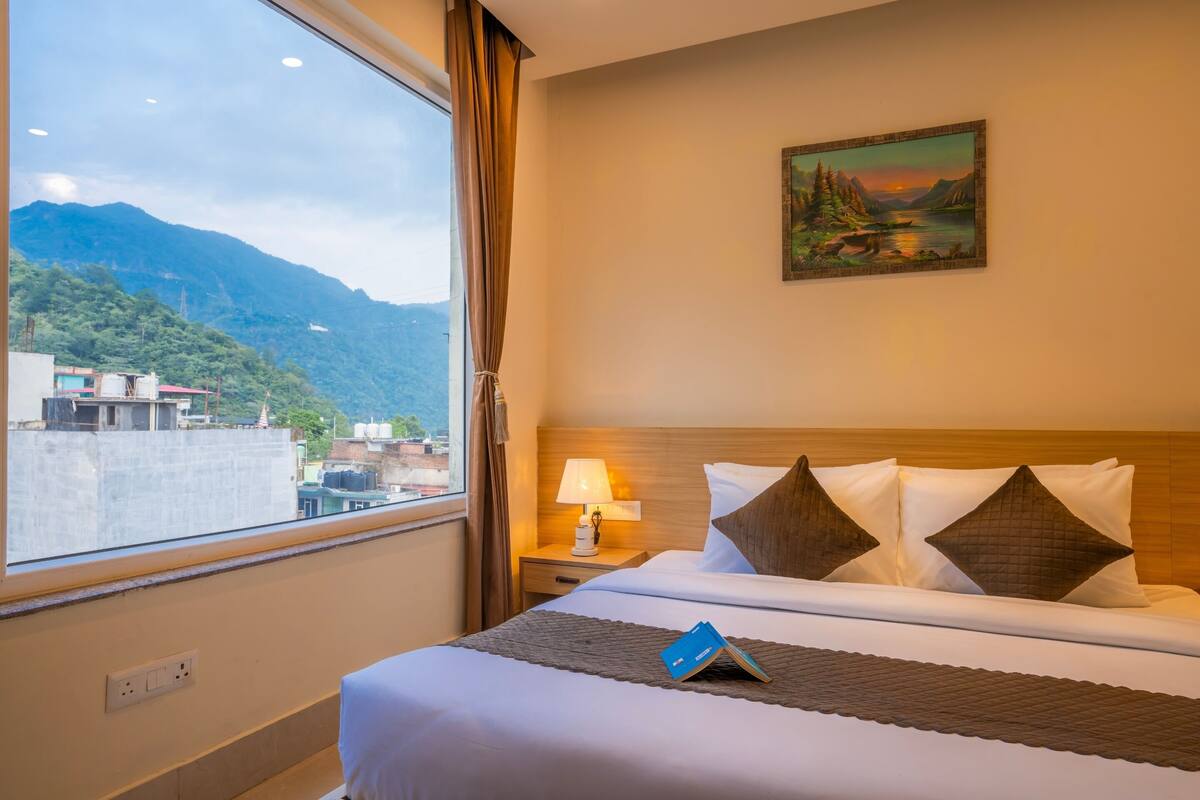 Una habitación de hotel con cama, mesita de noche con lámpara, un cuadro en la pared y ventana con vistas a montañas.