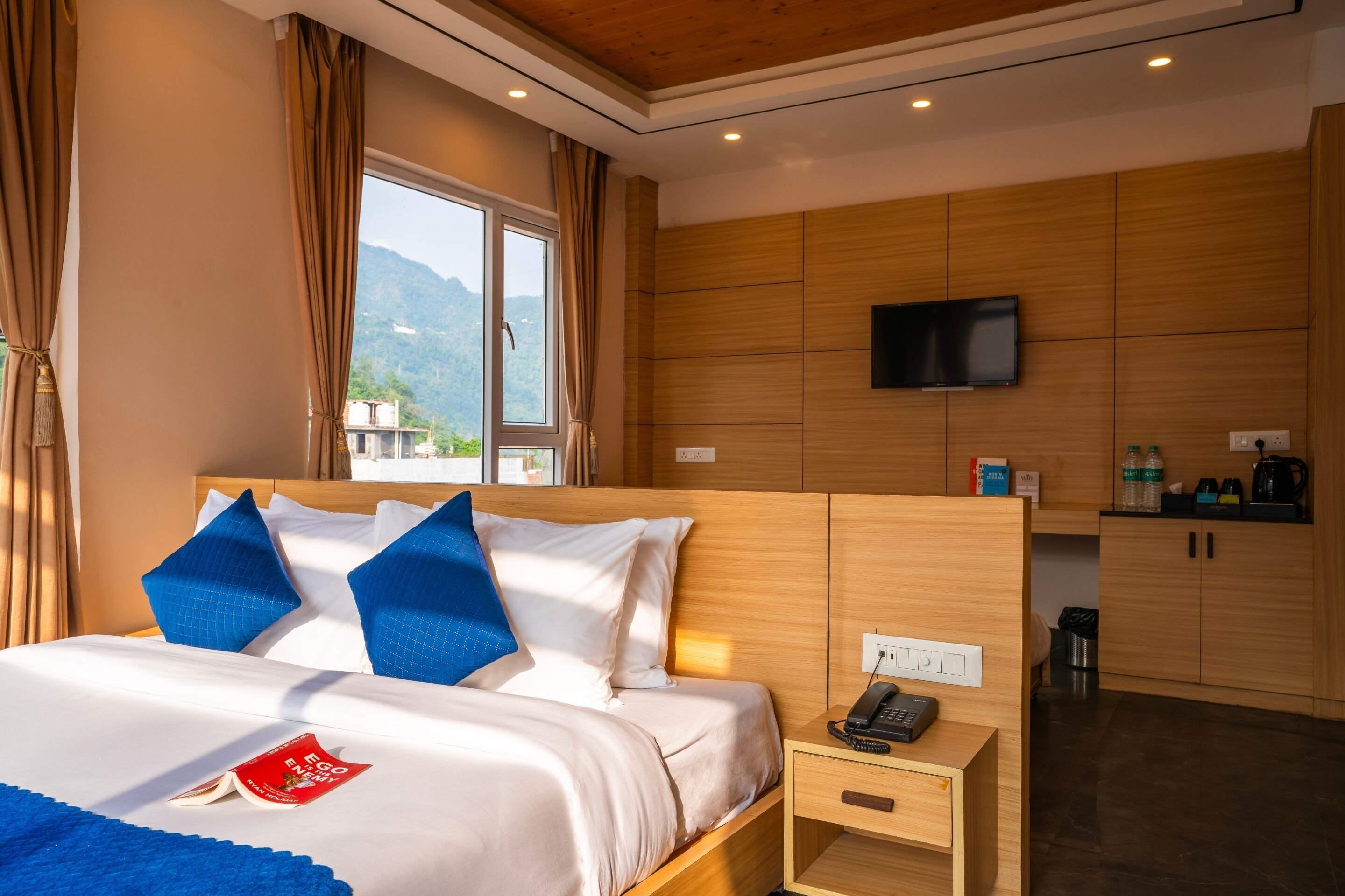 Quarto de hotel com uma cama, mesa de cabeceira com telefone, televisão na parede e vista para montanhas pela janela.