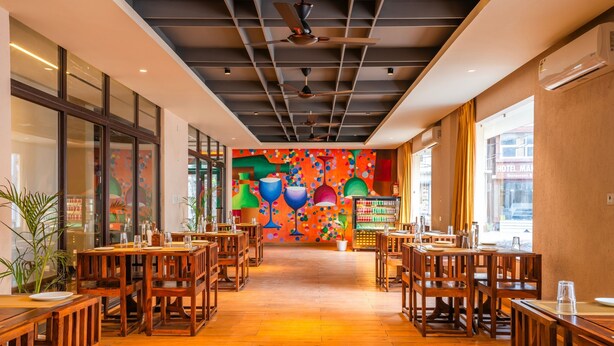 A restaurant with wooden tables and chairs, a colorful mural on the back wall, and large windows on the left side. - Moustache Select Rishikesh (Rishikesh)