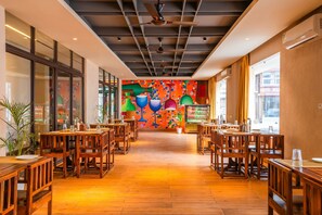 A restaurant with wooden tables and chairs, a colorful mural on the back wall, and large windows on the left side. - Moustache Select Rishikesh (Rishikesh)