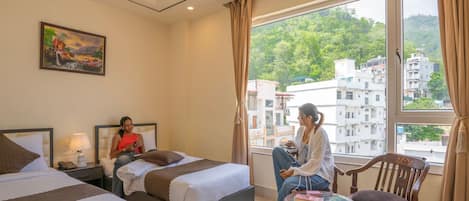 Habitación de hotel con dos camas, un área de descanso con una silla y una mesa, un cuadro en la pared y vista a edificios a través de la ventana.
