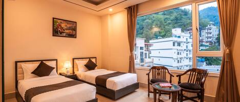 A hotel room with two beds, a small table with chairs, a painting on the wall, and a view of buildings through the window.