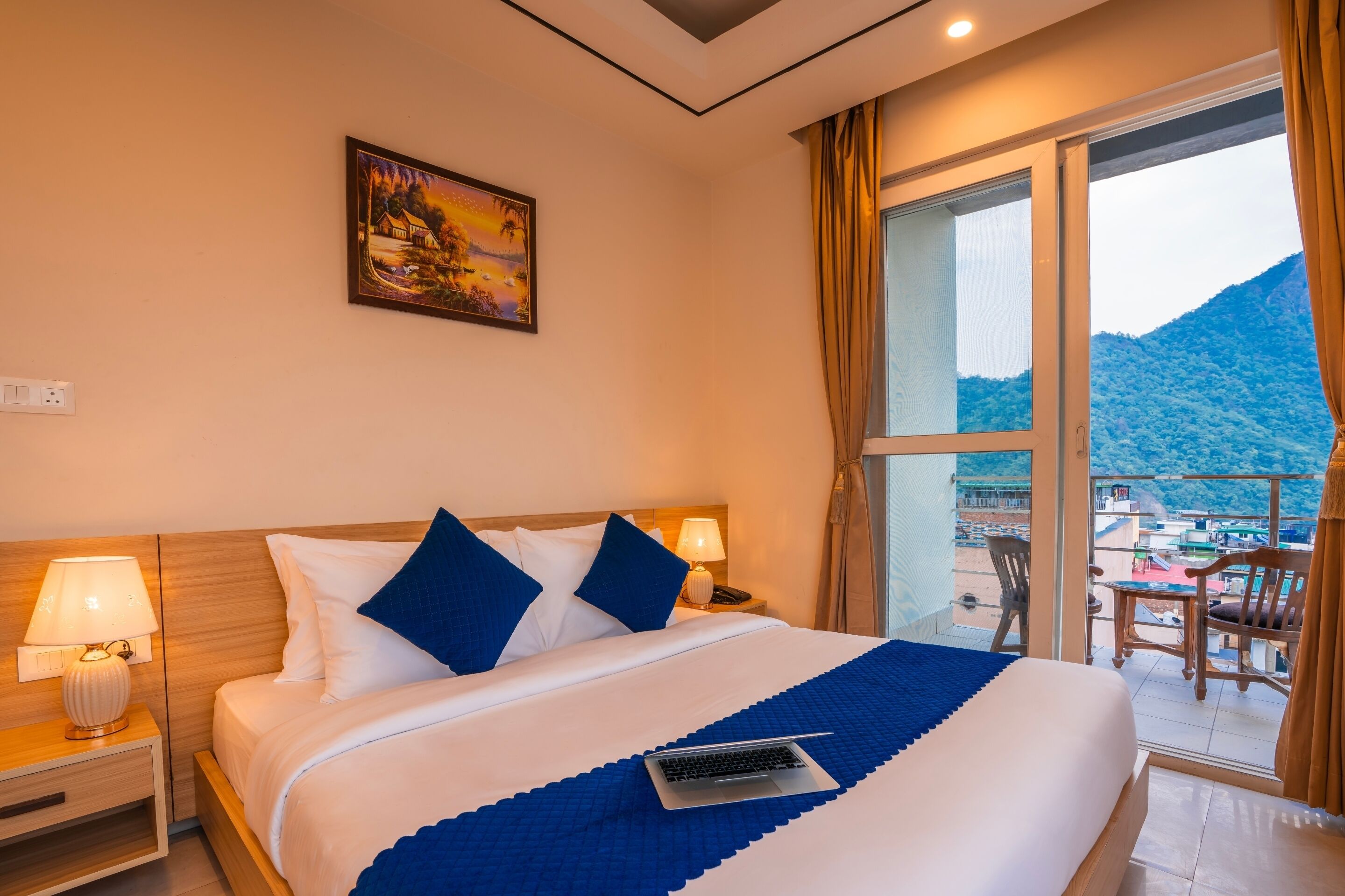 A hotel room with a bed, a laptop, a painting, and a balcony with a view of the mountains.