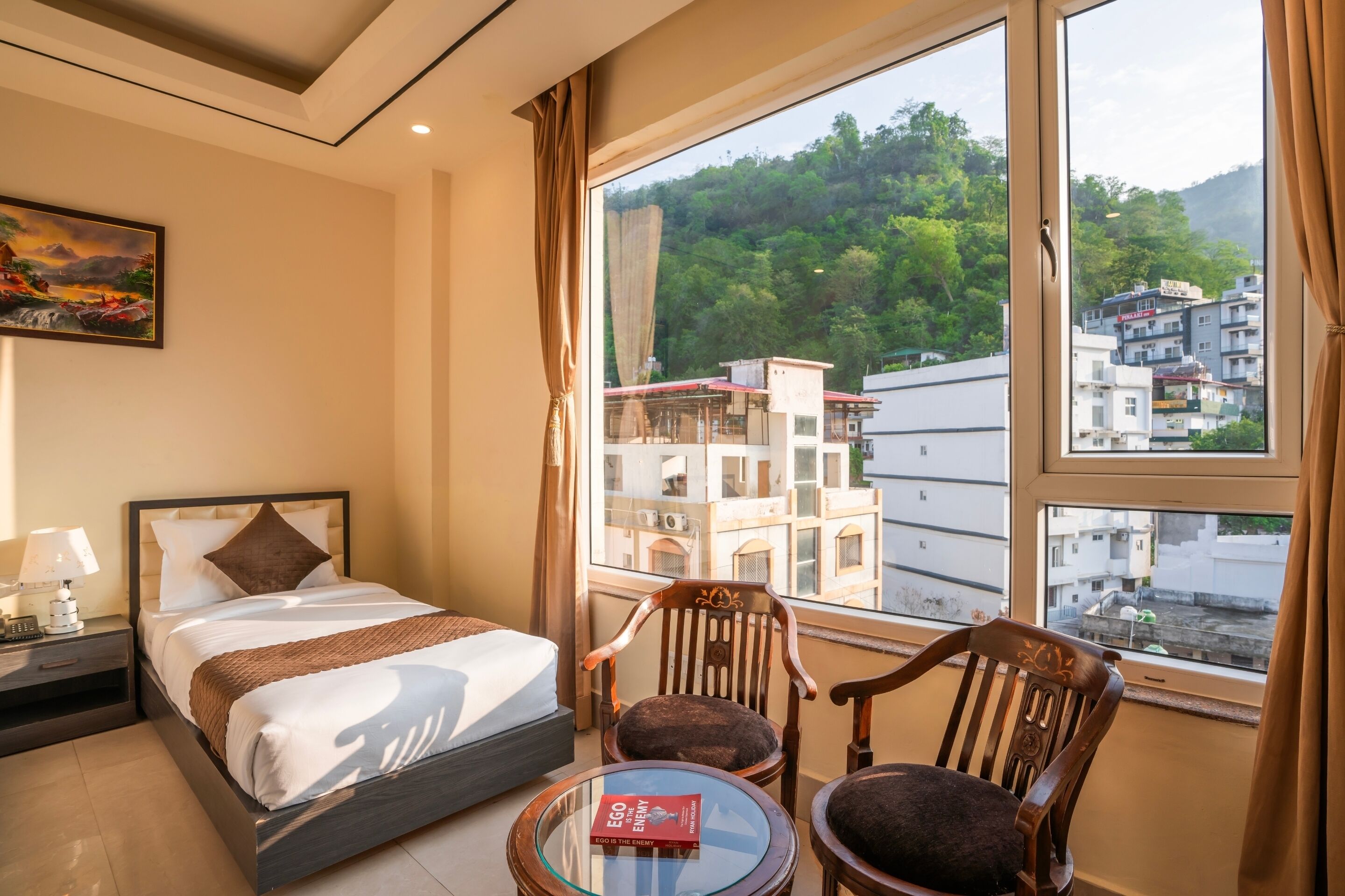 A hotel room with a bed, a chair, a small table, a painting on the wall, and a view of buildings and greenery outside.