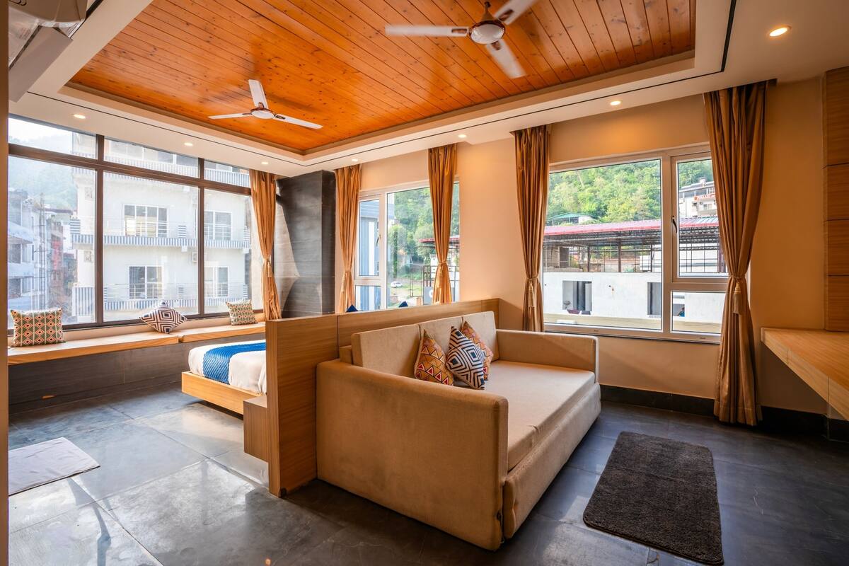 Una habitación moderna con sofá, cama y grandes ventanales con vistas a edificios y árboles.