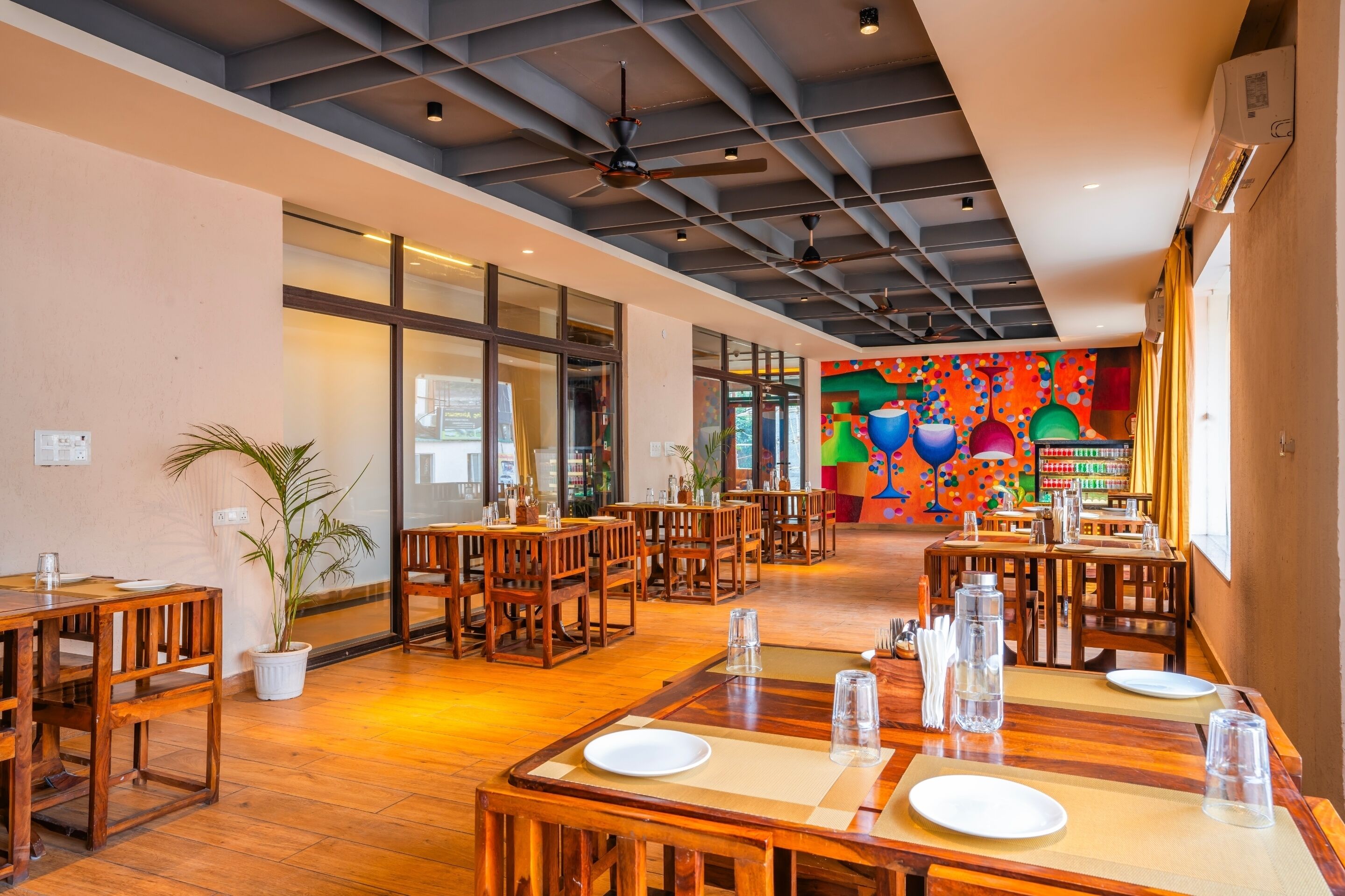 A spacious restaurant with wooden tables and chairs, large windows, and a colorful mural on the back wall.