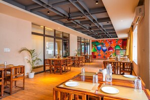 A spacious restaurant with wooden tables and chairs, large windows, and a colorful mural on the back wall.