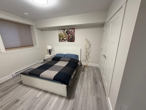 A bedroom with a bed, bedside table, lamp, artwork, wardrobe, and a window with blinds.