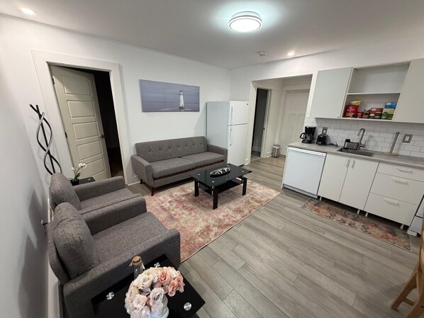 A modern living room with a sofa, armchairs, a coffee table, and a rug. There is a kitchen area with cabinets and a countertop.