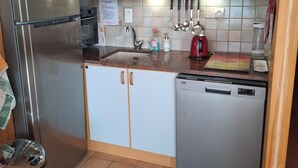A compact kitchen with a stainless steel refrigerator, a dishwasher, and a sink with a faucet. There are wooden cabinets with glass doors, a hanging light fixture, and a tiled backsplash. - 3-room apartment 55 m2, terrace 15 m2, breathtaking view, sleeps 4, air-conditioned, WIFI (Sainte-Croix-du-Verdon)